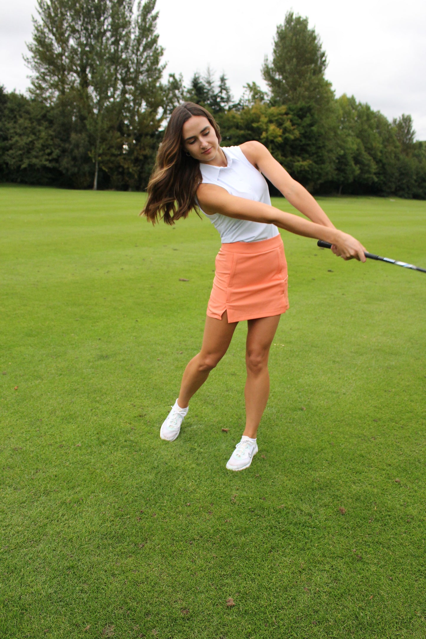 AWGO Golf Coral Skort on female golfer Women's high-waisted golf skort in coral Performance golf skirt with stretchy fabric Close-up of pocketed golf skort in coral Female golfer wearing AWGO Golf Coral Skort Stylish women's golf apparel in coral Sporty golf outfit with high-waisted skort Golf skort with convenient pockets AWGO Golf Coral Skort for female golfers Coral golf skort for performance and style Woman playing golf in Golf Skort Woman Swinging Golf Club Woman making golf shot in golf skirt